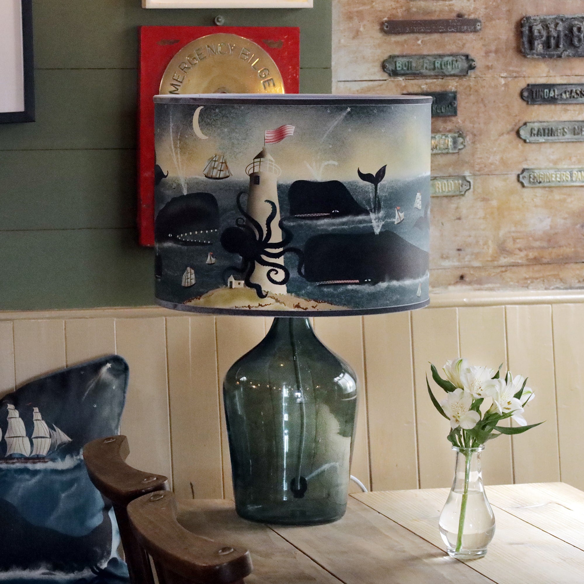 A large glass lamp with a nautical-themed lampshade sits on a wooden table. The lampshade features an ocean scene with a lighthouse being enveloped by a giant octopus, several whales swimming nearby, ships with sails, and a crescent moon above. A small vase of white flowers is placed next to the lamp. The backdrop includes a rustic wall with metal signs and a red "Emergency Bilge" plaque, while a cushioned chair with a maritime-patterned pillow sits nearby, reinforcing the sea-inspired ambiance.