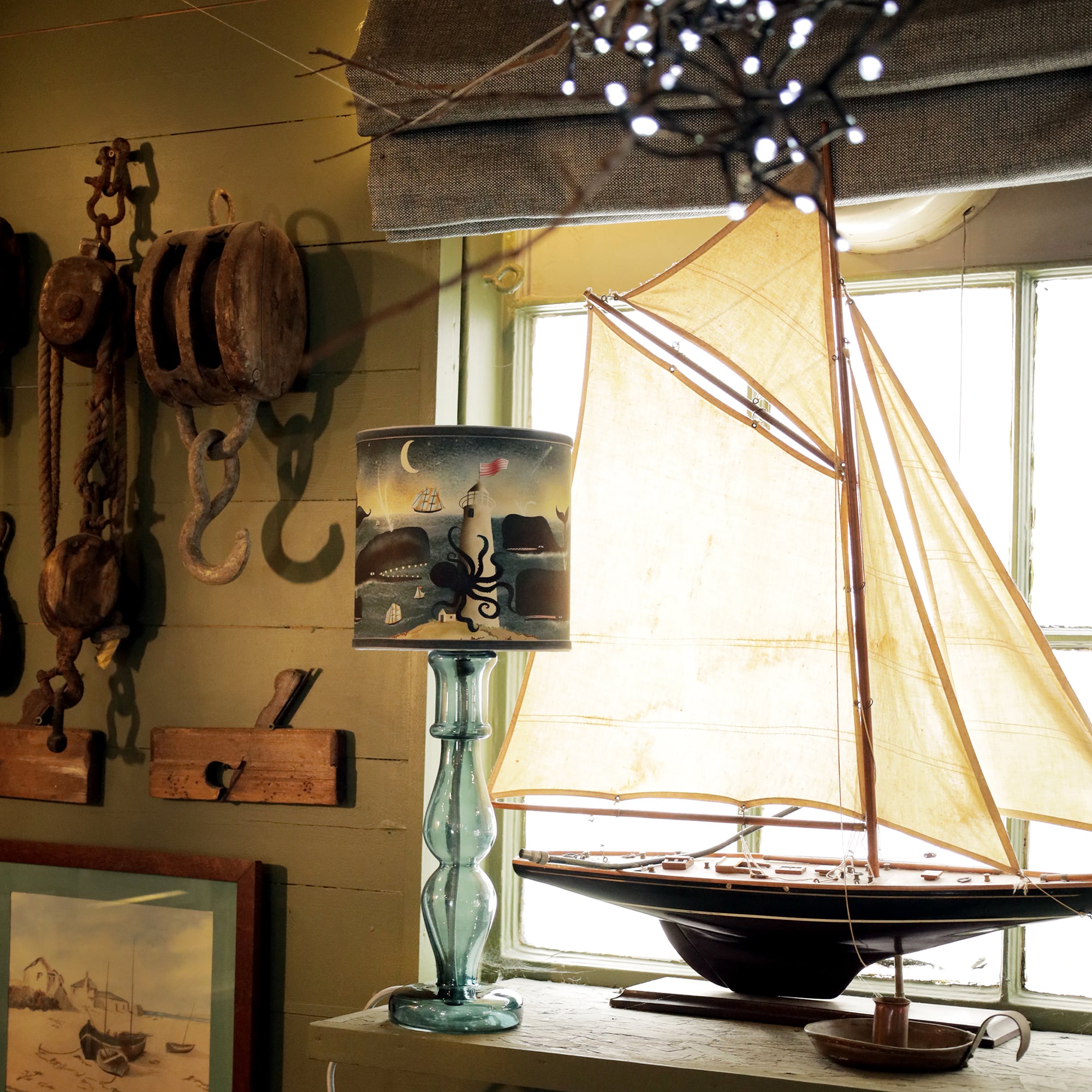 A cozy, nautical-themed interior featuring a glass table lamp with a marine-inspired lampshade. The lampshade depicts a playful ocean scene with whales swimming around a lighthouse, accompanied by sailboats, an octopus, and a crescent moon. The lamp sits next to a large model sailboat with cream-colored sails, placed on a windowsill. The surrounding decor includes rustic wooden pulleys and maritime tools hanging on the wall, adding to the room’s coastal, vintage aesthetic. Soft lighting enhances the warm, s