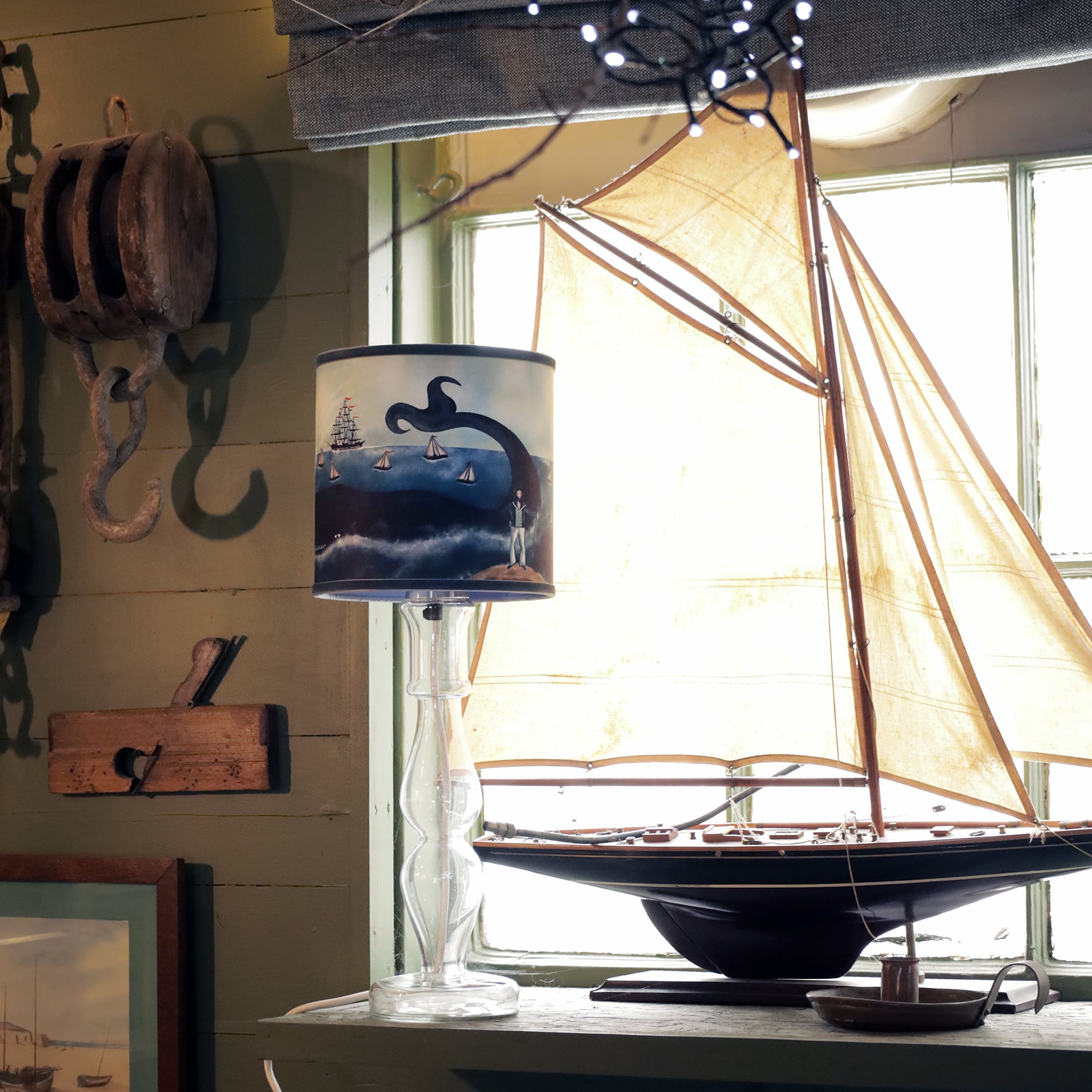 A cozy, vintage-style room featuring nautical decor. A small model sailboat with tan sails sits on a wooden shelf beside a transparent glass lamp. The lampshade depicts a whimsical scene of a sea monster rising from the water near small sailboats. Nautical elements, like a pulley and a ship hook, hang on the green wooden wall behind, completing the maritime theme. Warm light streams through the window, casting a nostalgic and charming atmosphere.