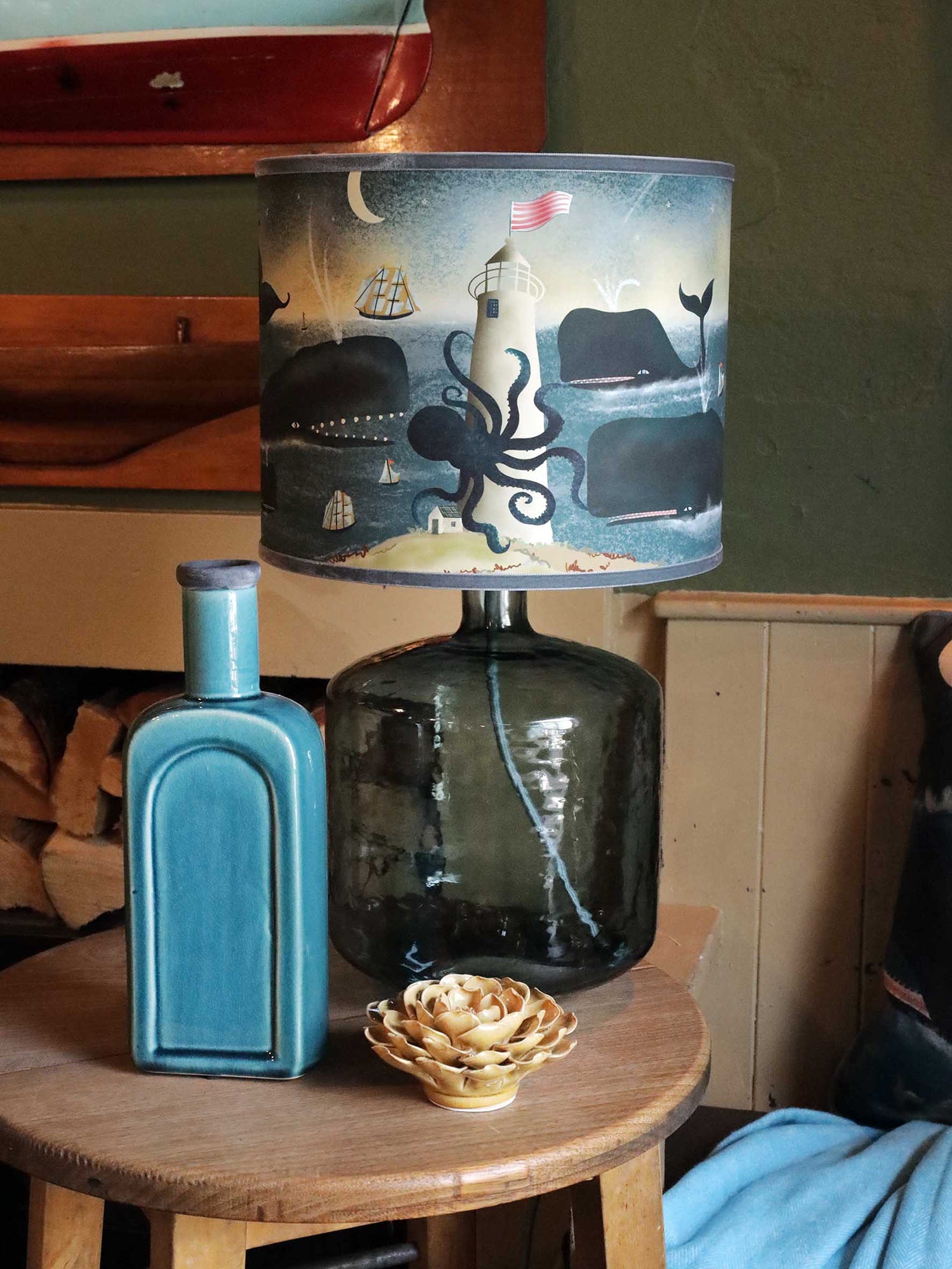 A cozy, rustic room featuring a round wooden side table with a large glass lamp and a marine-themed lampshade. The lampshade depicts a whimsical ocean scene with whales swimming around a lighthouse, accompanied by sailboats, an octopus, and a crescent moon in a tranquil night sky. On the table next to the lamp, there is a teal ceramic bottle and a decorative ceramic flower, adding a touch of coastal charm. The background includes a wall with warm wooden tones and a small stack of firewood, contributing to t