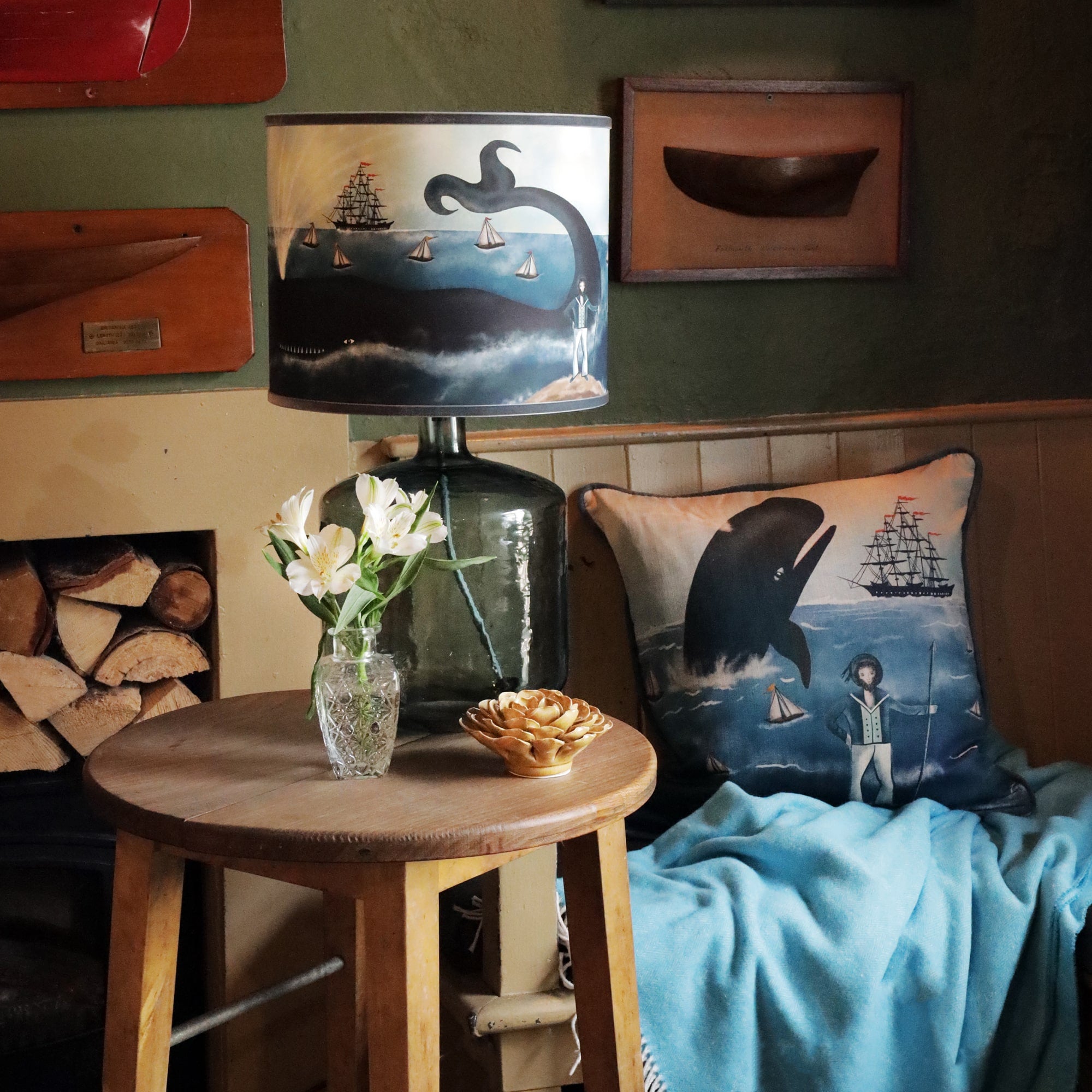  A cozy, nautical-themed corner featuring a wooden side table with a large glass lamp and a maritime lampshade. The lampshade showcases a whimsical ocean scene with a whale, sailboats, a tall ship, and a captain on the shore. On the table, there is a vase of white flowers and a ceramic decorative piece. Beside the table, a matching cushion with the same nautical design rests on a chair draped with a blue blanket. The room is decorated with wooden nautical artifacts and warm tones, evoking a charming seaside