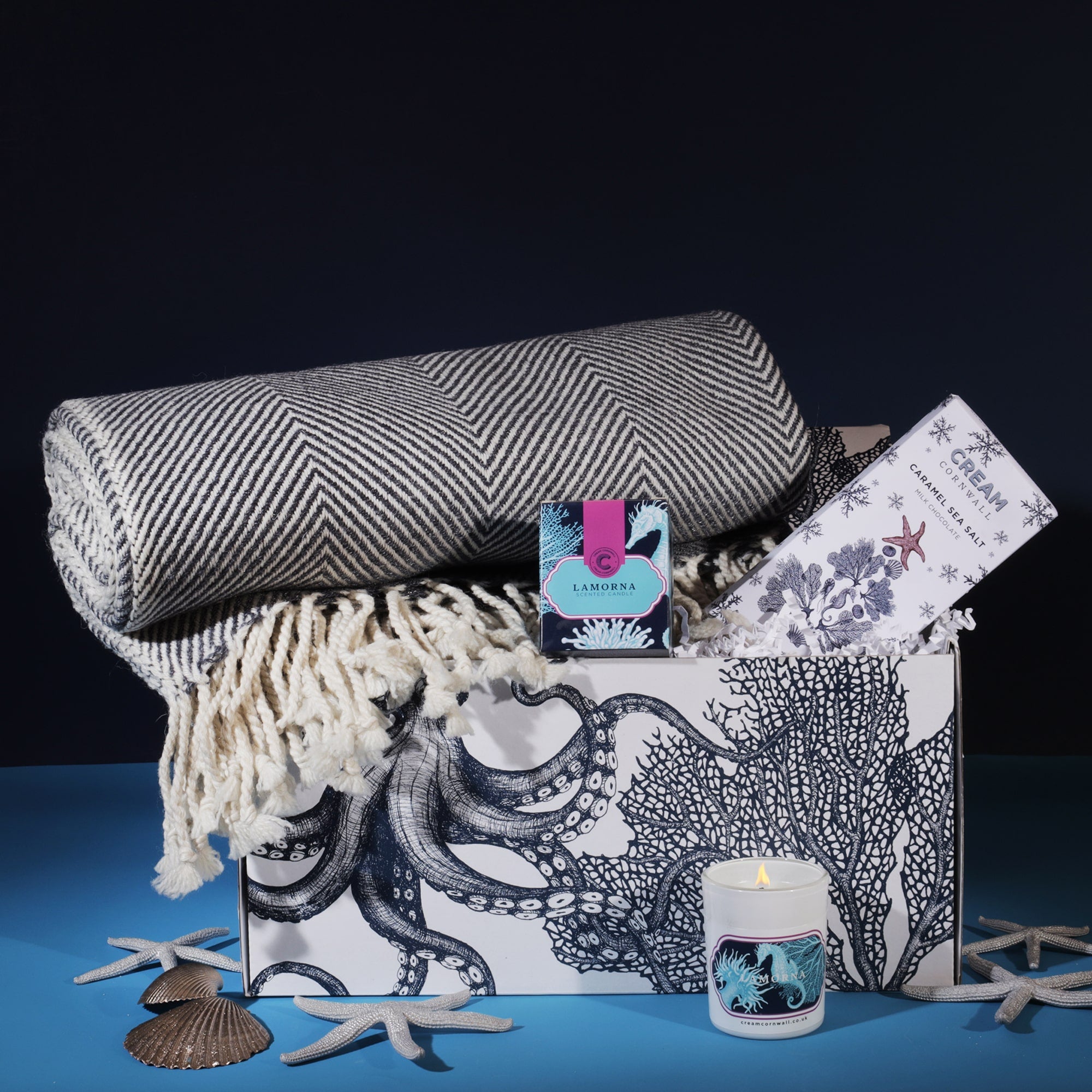 A Cream Cornwall gift set featuring a rolled-up herringbone-patterned throw blanket, a bar of caramel sea salt chocolate, a Lamorna scented candle, and a matching box. The items are arranged in a gift box adorned with intricate octopus and coral illustrations. Decorative seashells and starfish are scattered around the set, enhancing the coastal theme against a dark blue background.