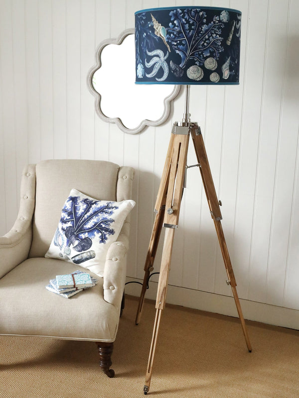 Wooden Tripod with a Rockpool Navy lampshade next to a chair .On the chair is a white matching Rockpool cushion with notebooks placed on the chair.Behind on the wall is a white wave mirror
