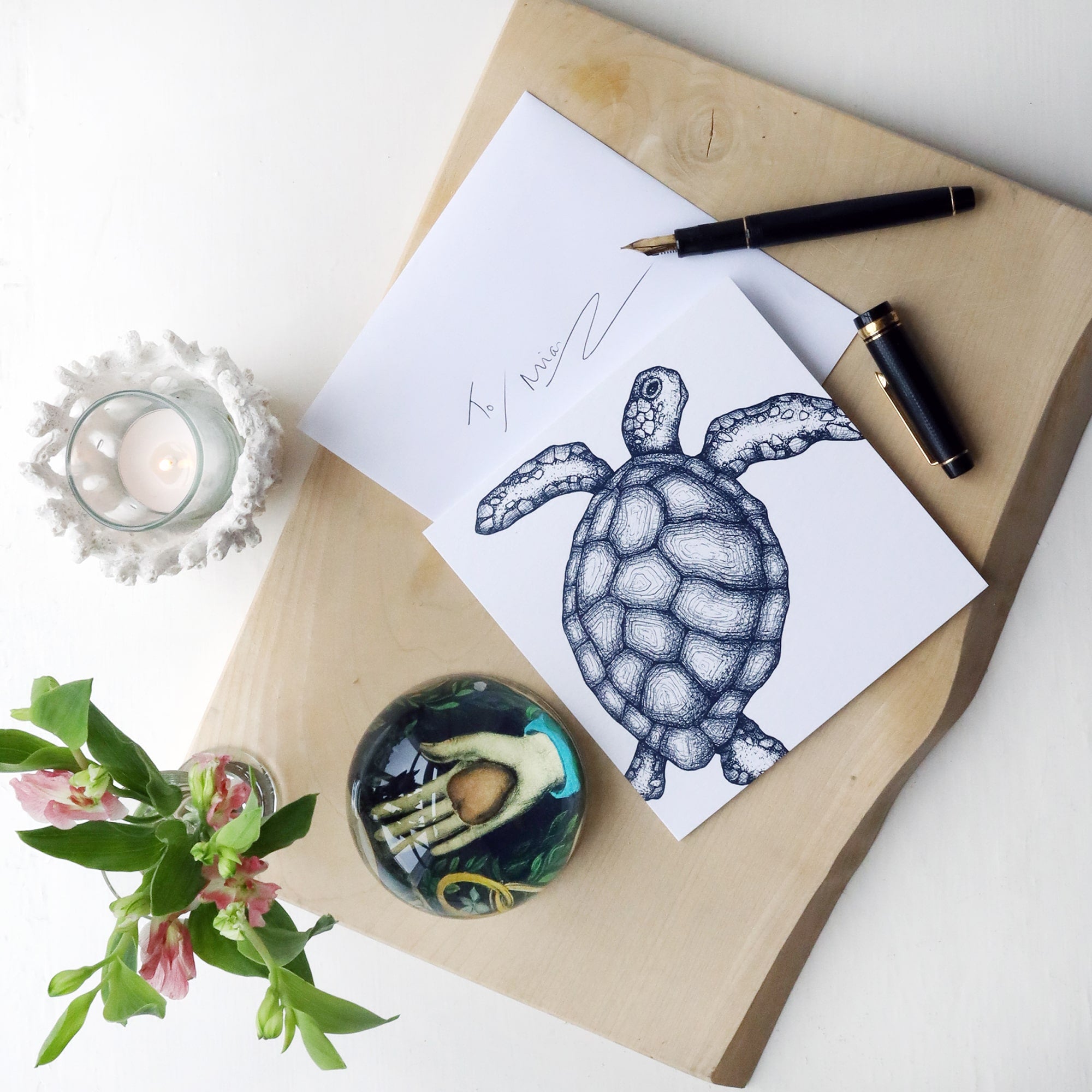 White greeting card with navy illustrated turtle on, lying on a wooden block with fountain pen, paperweight and flowers and hand written envelope.