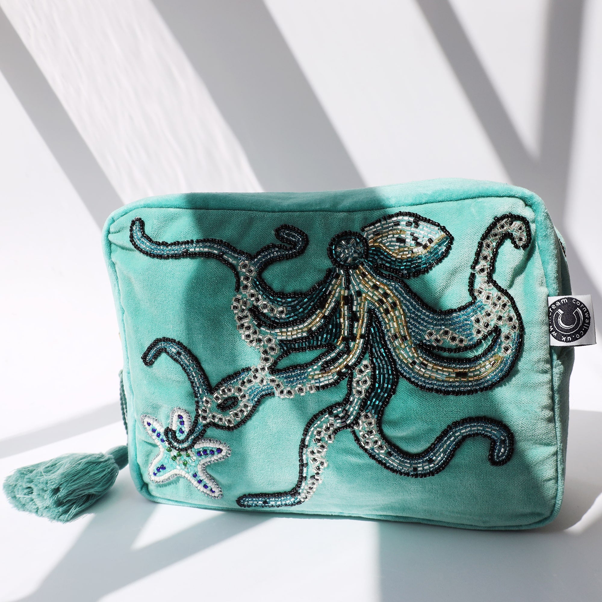 Beaded aqua velvet wash bag with octopus and starfish decoration on a white background with the sun casting shadows over it.