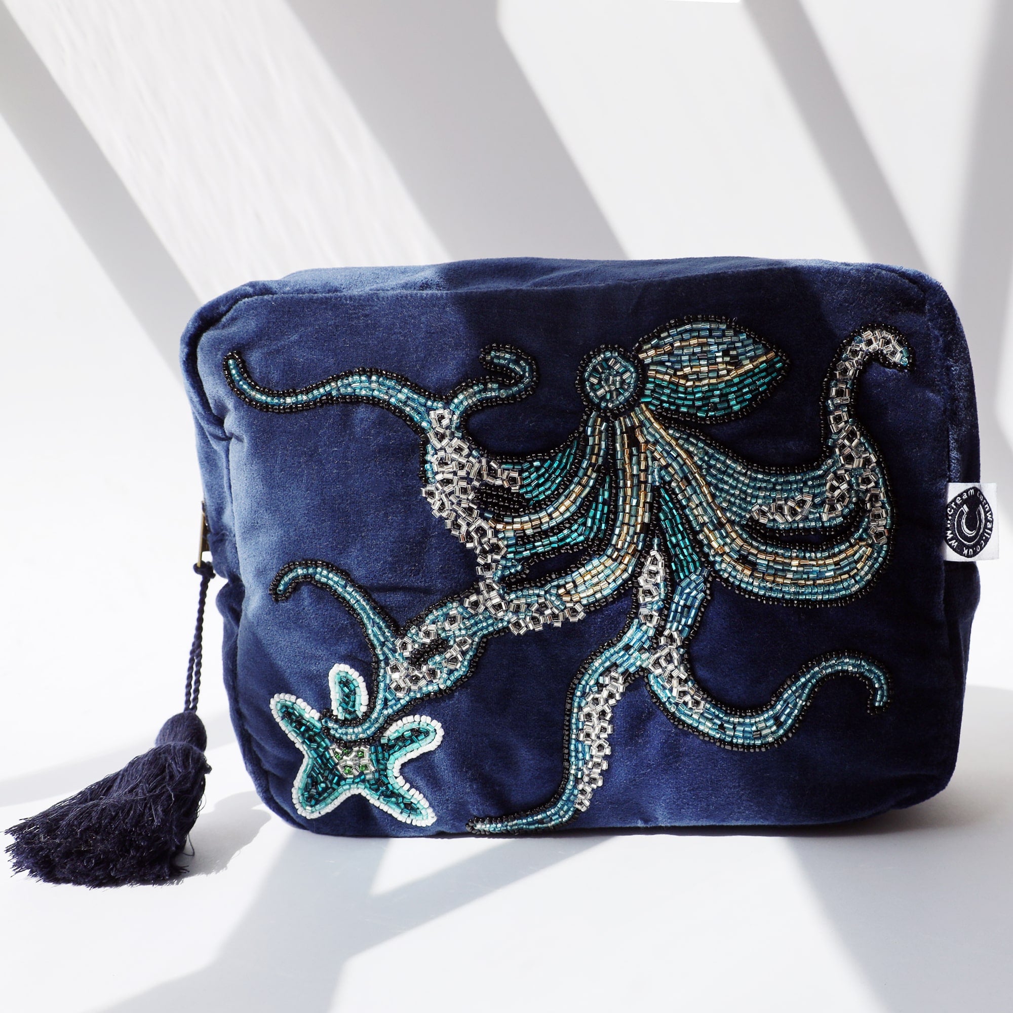 Beaded wash bag in navy velvet with octopus and starfish on the front. The bag has a large cotton navy tassel hanging from the zip and is sitting on a white background with the sun casting shadows across it.