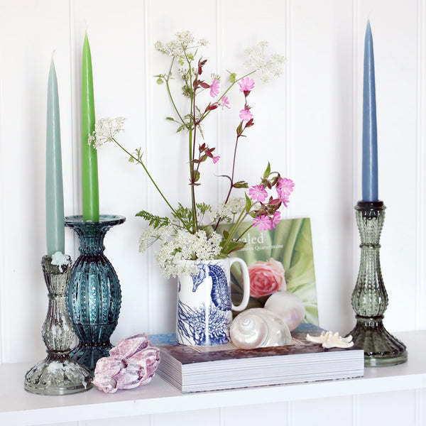 3 and green blue toned taper dinner candles win glass candle sticks, sitting on a white shelf with books and a blue and white seahorse jug with wild flowers