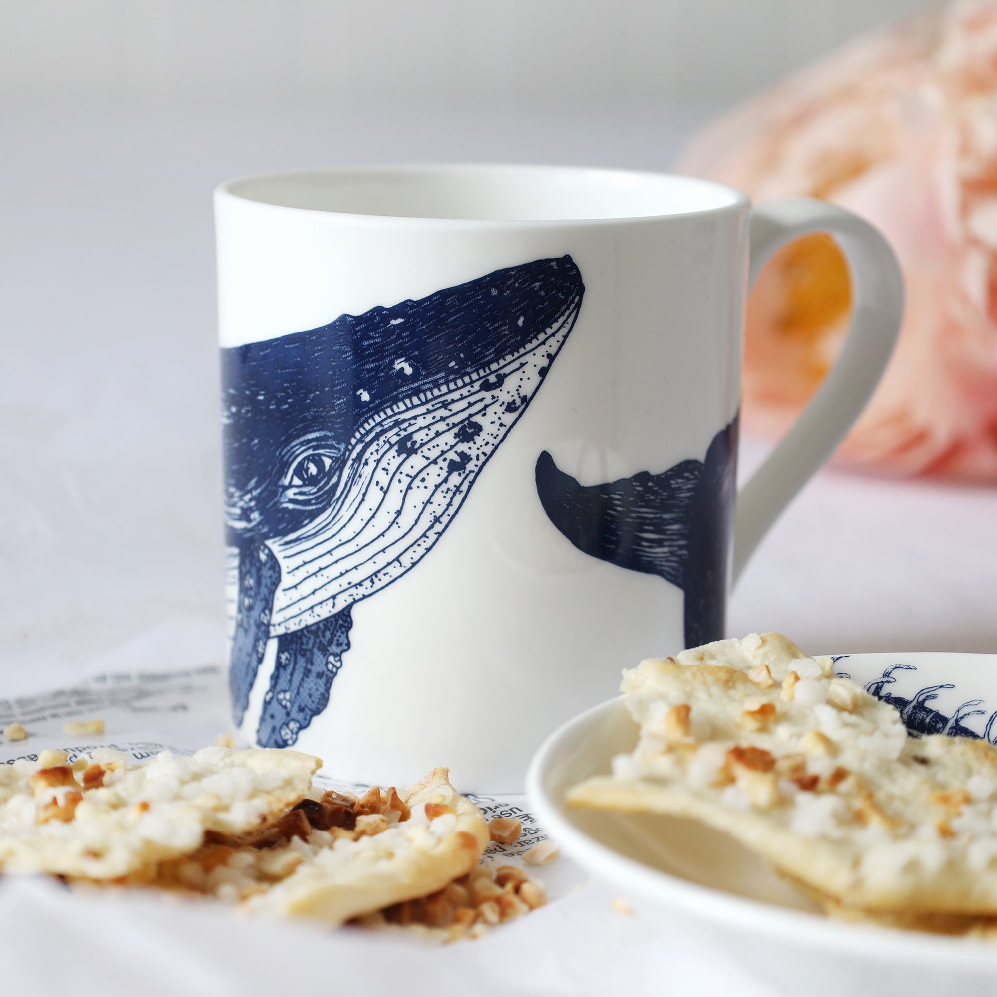 White mug with dark blue humpback whale illustration sitting on a table with a plate & biscuits and flowers in the background.