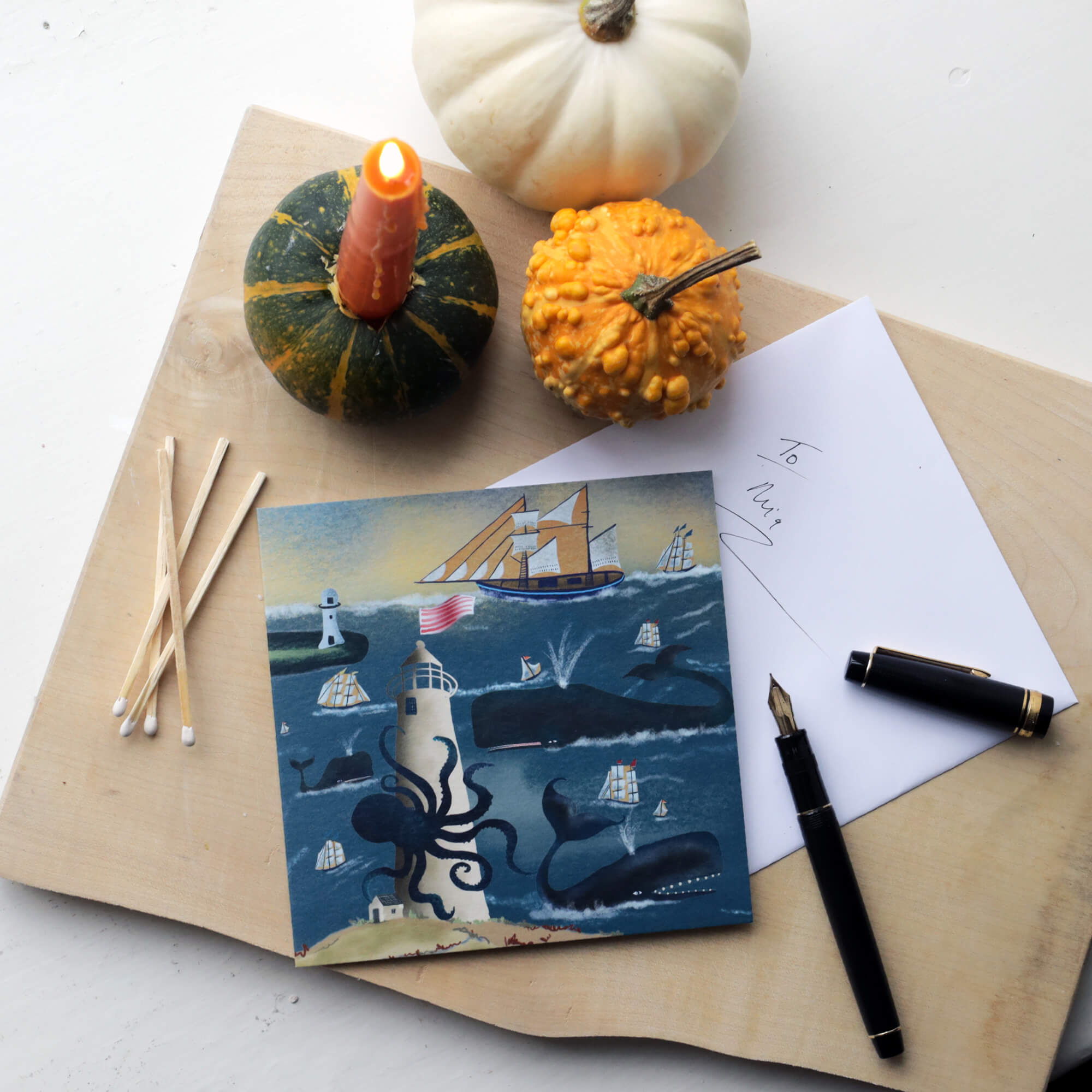 A flat lay display featuring a nautical-themed greeting card with whales, ships, and a lighthouse. The card is placed on a wooden surface alongside an envelope, a fountain pen, and a note addressed 'To Mum.' Surrounding the card are autumnal decorations, including a lit candle inserted into a small green pumpkin, a white pumpkin, a yellow gourd, and several matches.