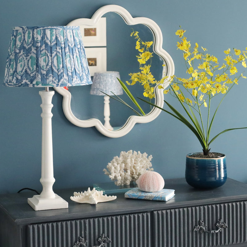 White Lacquered wavey edged shaped mirror on the wall.In front is a sideboard with an yellow flowered plant,some shells,a notebook and one of our white lamp bases and a pleated lampshade