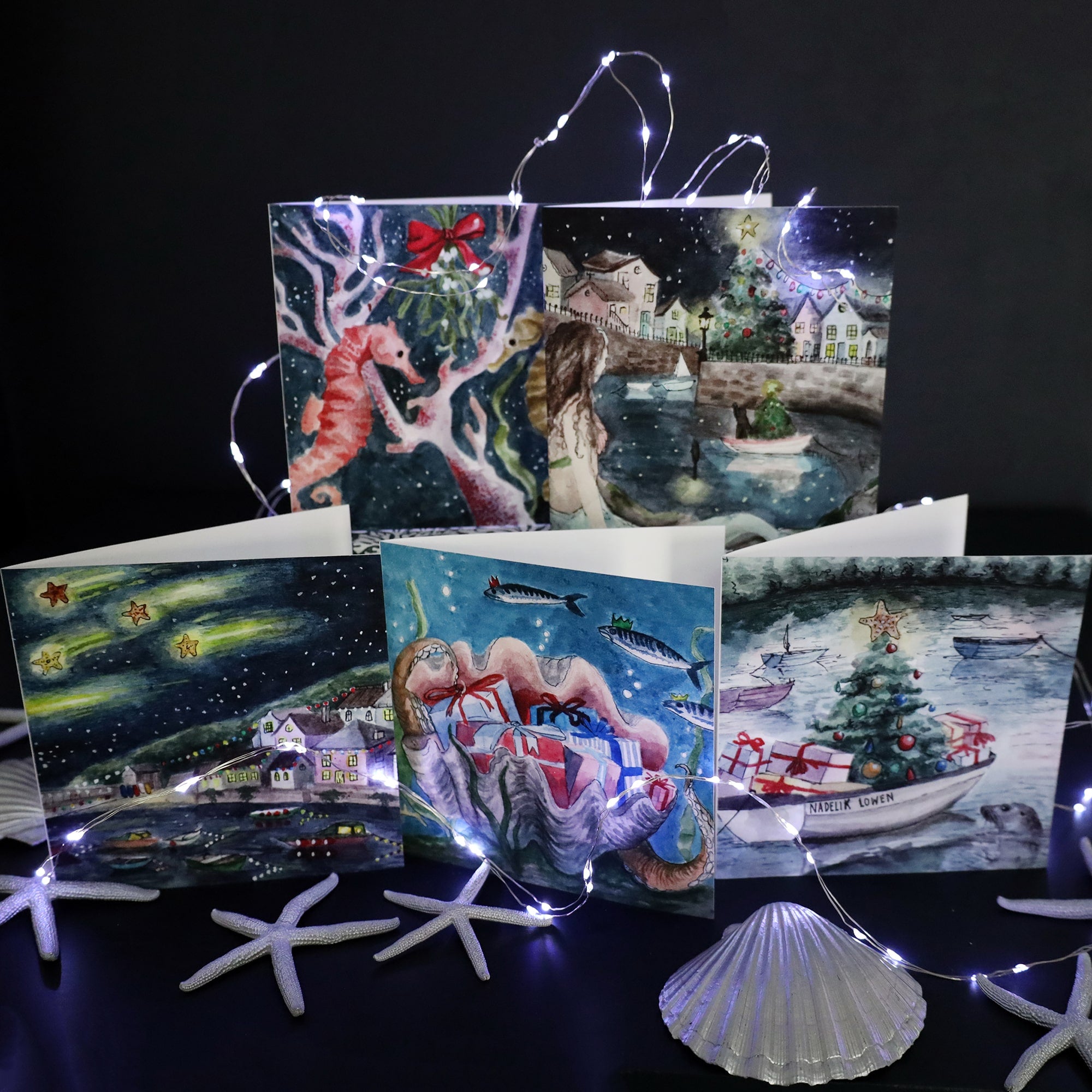 A display of five Cream Cornwall Christmas cards arranged against a dark background, illuminated by string lights. The cards feature watercolor illustrations of festive coastal scenes, including seahorses around coral with a red ribbon, a seaside town at night, a large seashell filled with presents, and a boat decorated with a Christmas tree. One card shows a boat with 'Nadelik Lowen' (Merry Christmas in Cornish) written on it, with a seal peeking out of the water.