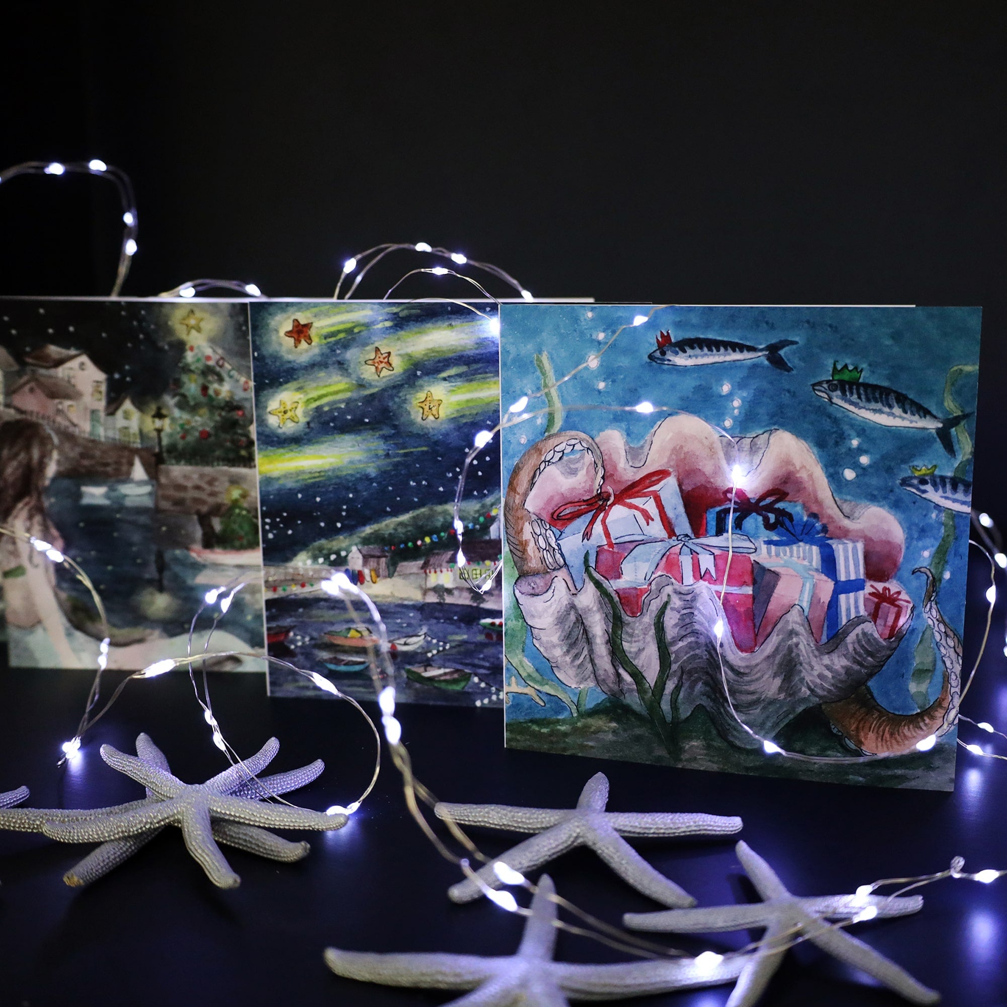 Three Cream Cornwall Christmas cards displayed with string lights and starfish decorations. The cards feature festive coastal scenes, including a seaside town with a Christmas tree, a starry night sky over a harbor, and a large seashell filled with wrapped presents, surrounded by fish wearing crowns. The string lights and starfish accents add a whimsical, nautical touch to the holiday theme.