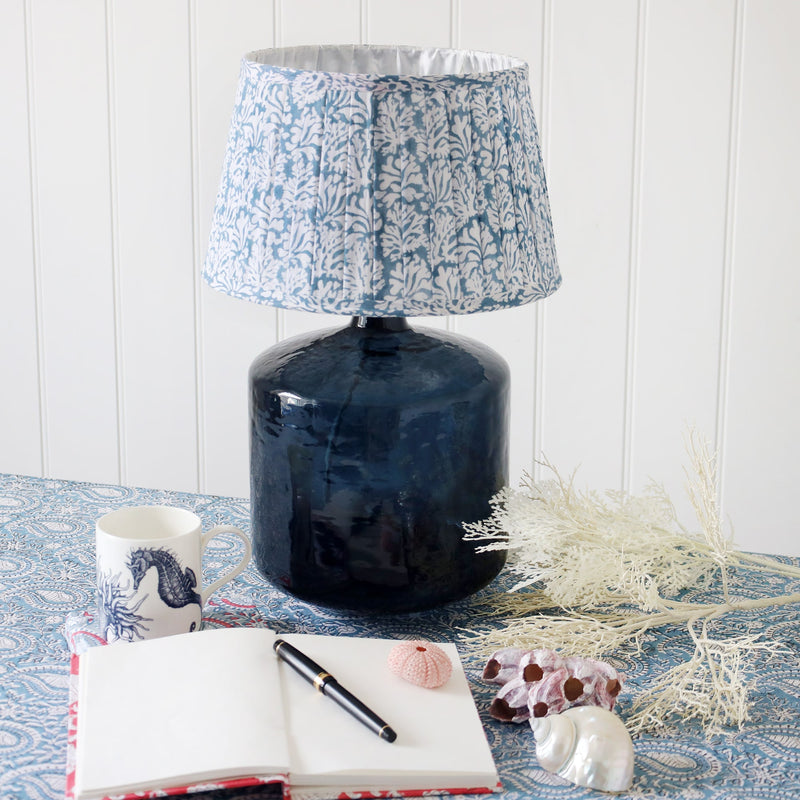 Medium Coastal Blue pleated lampshade in hand blocked print in blue and white on a zennor glass lampbase on a table.Also on the table is an open notebook with a pen and a seahorse mug,also shells and coral on the table