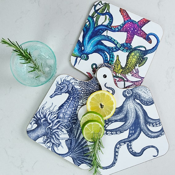 Seahorse mini chopping board placed on a table with lemon and limes next to a glass filled with ice.The chopping board is on top of other different chopping boards