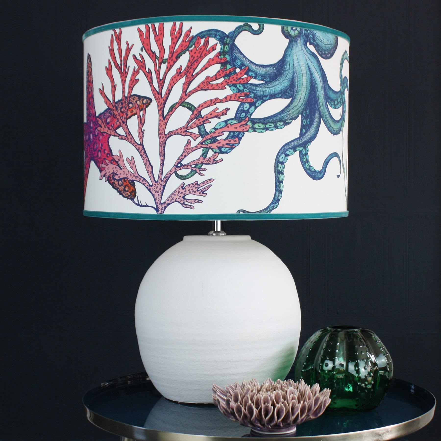 Rainbow Reef White Shade With Octopus,Seahorse,Starfish and Seaweed Design in bright colours on a white lampbase on a table with a lilac coral and a green glass vase.