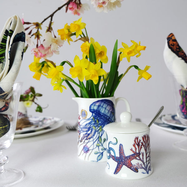 Bone China Jellyfish Reef Jug and Jam pot with hand drawn illustrations.In the jugs are bright yellow daffodils in the background are matching reef tableware 