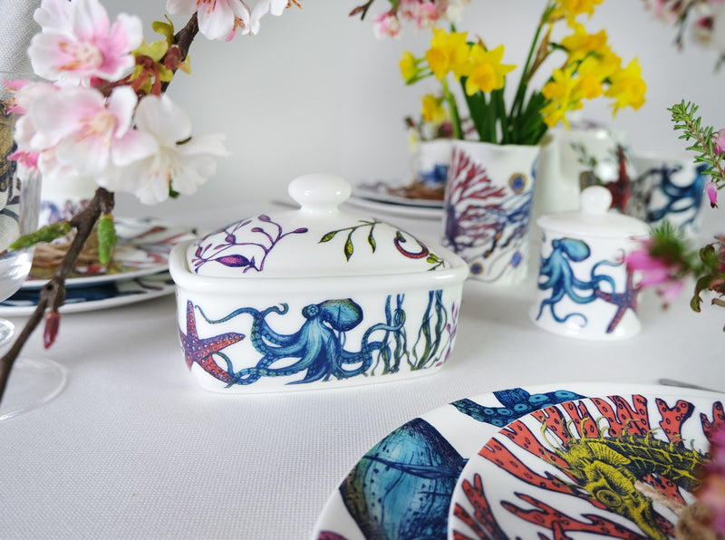 Butter dish in our brightly coloured Reef range,with Octopus,seaweed,seahorses and other sea themed designs all over the base and the lid.It is set on a table surrounded by other pieces from the Reef range and lots of bright flowers in vases are out of focus in the background
