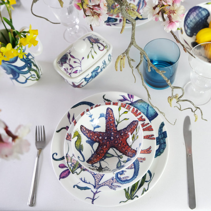 Bowl in Bone China in our Reef range in bright colours in the Starfish design in a stack with side plate and dinner plate placed on a white tablecloth with cutlery. On the table are matching butter dish,jugs and other bowls with a couple of glasses in between 