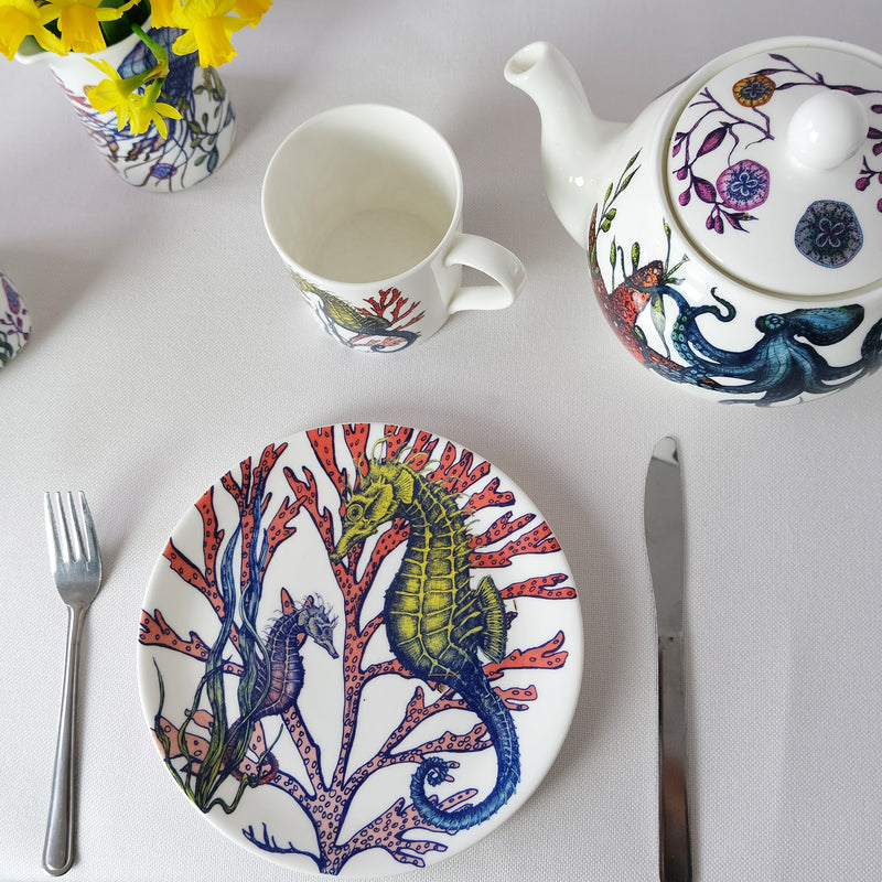 Bone China White plate with hand drawn illustrations of our Seahorse Reef design on a side plate in beautiful bright colours.Next to it is a Reef Teapot and a reef mug