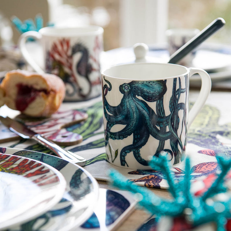 Close up of  the Reef Octopus Mug on a Reef table runner with Reef plates in the foreground.Behind you can see a jam donut on a Reef chopping board and a seahorse mug in the distance