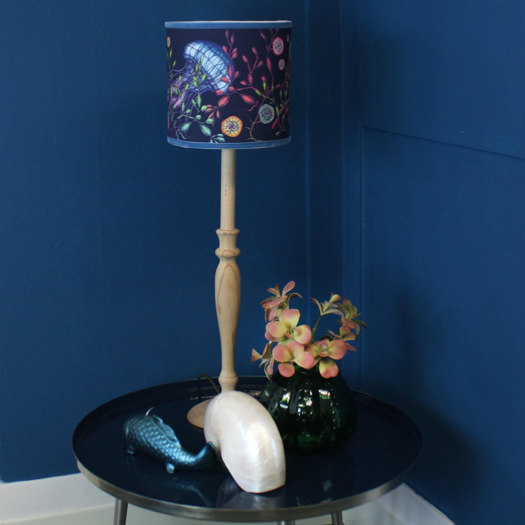 Canyons Reef Navy Shade With Jellyfish Design And Velvet Trim on a wooden lampbase placed on a blue table with a blue fish candle,large shell and a small vase with flowers