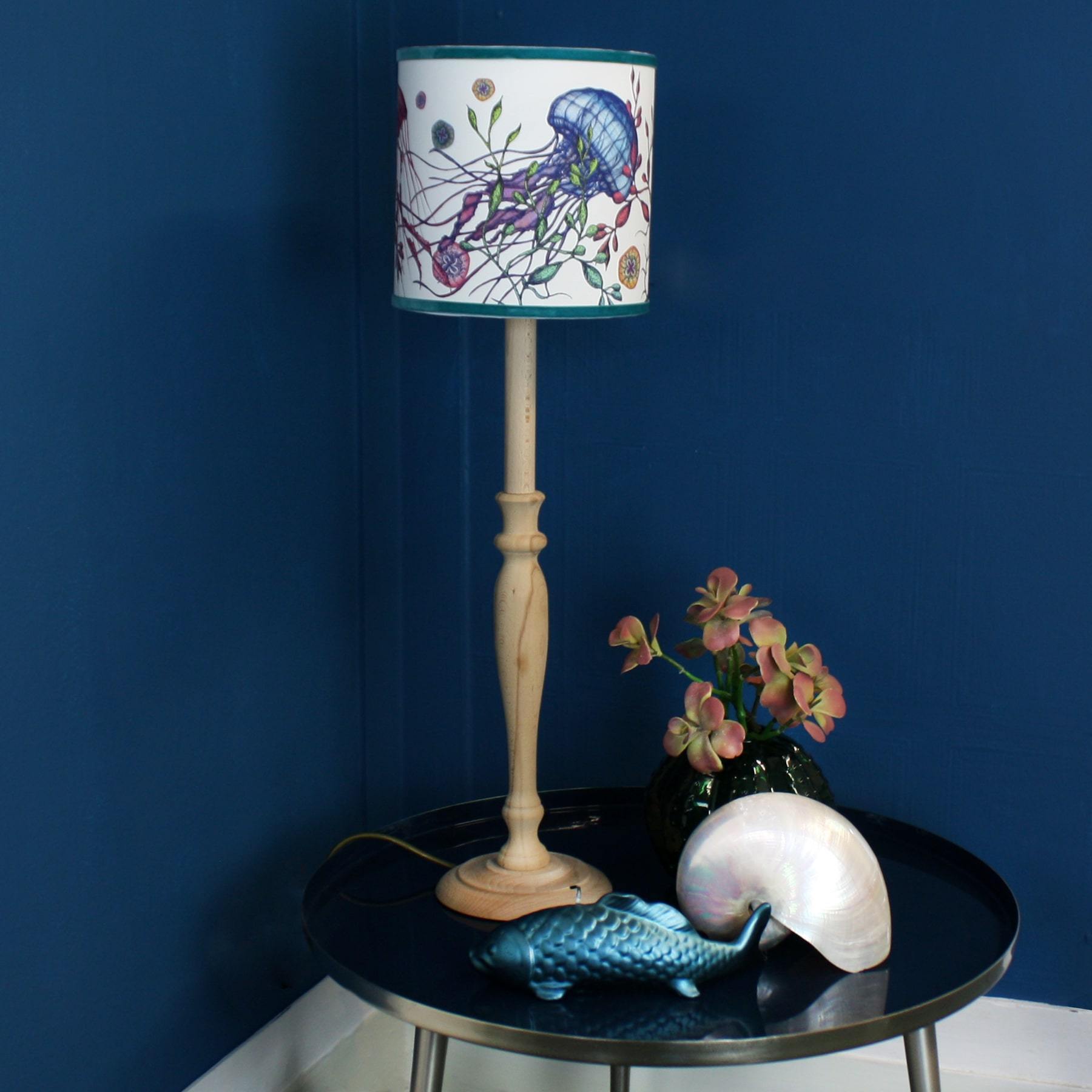 Canyons Reef White Lampshade With Jellyfish Design on a wooden lampbase on a small navy table with a fish candle,shell and a small vase with flowers on the table.
