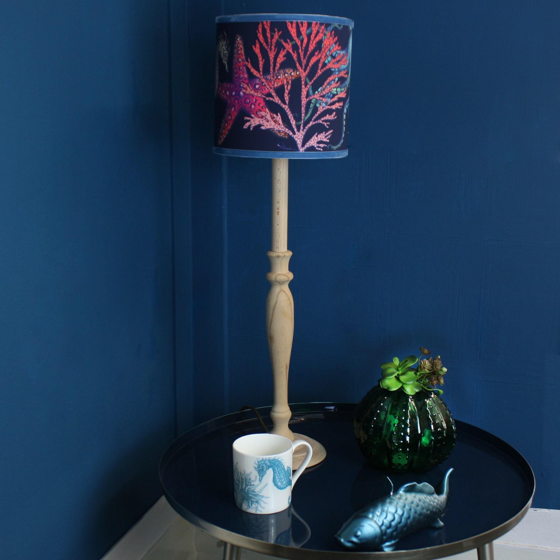 Rainbow Reef Navy Shade With Octopus,Seahorse,Starfish and Seaweed Design in bright colours on a wooden lampbase placed on a table with a fish candle,mug and a vase with flowers.