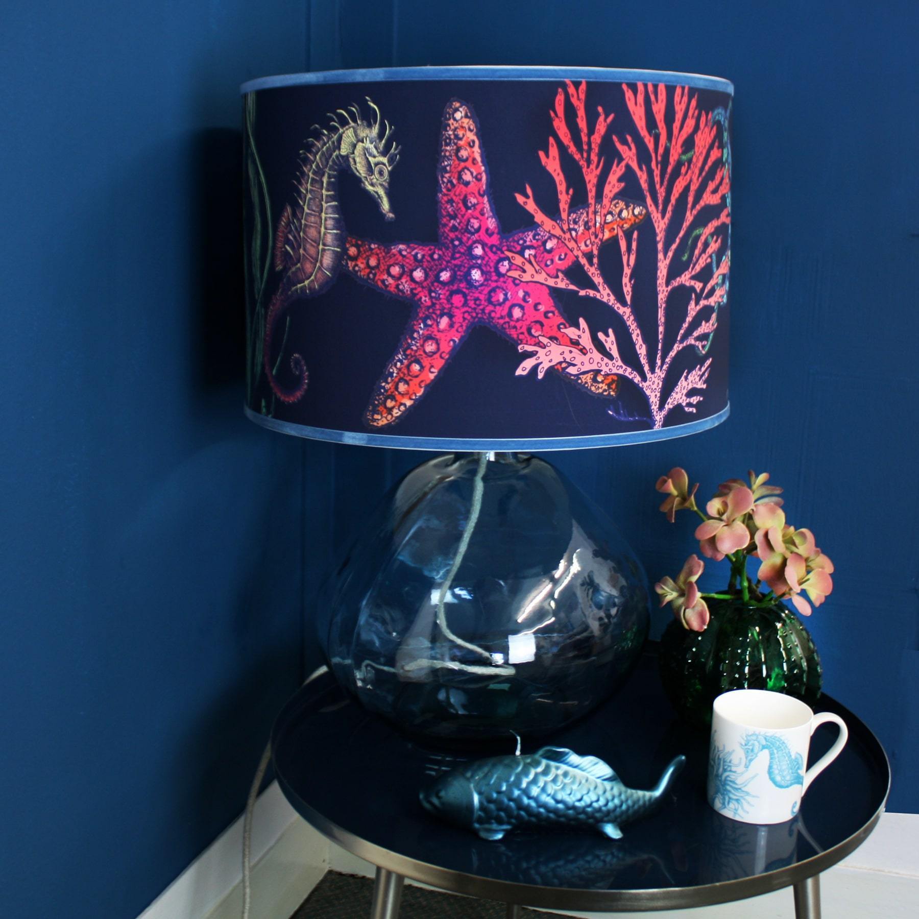 Rainbow Reef Navy Shade With Octopus,Seahorse,Starfish and Seaweed Design in bright colours on a blue glass lampshade placed on a side table with a fish candle,mug and a vase with flowers.