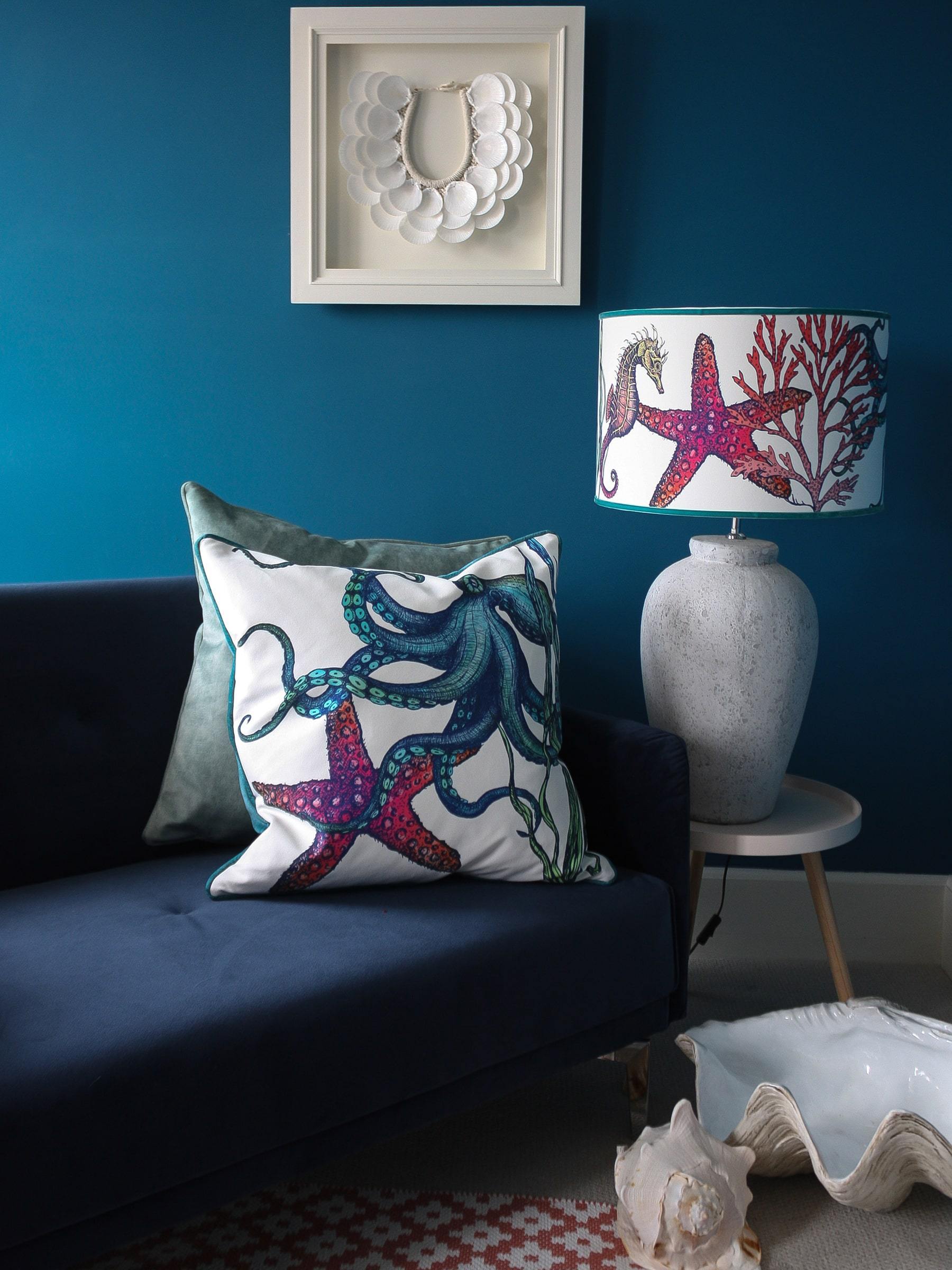 Rainbow Reef White Shade With Octopus,Seahorse,Starfish and Seaweed Design in bright colours on a white lampbase next to a navy sofa covered in our Reef cushions.Behind on the wall is a scallop art piece and in front of the sofa is a footstool and several shells on the floor.