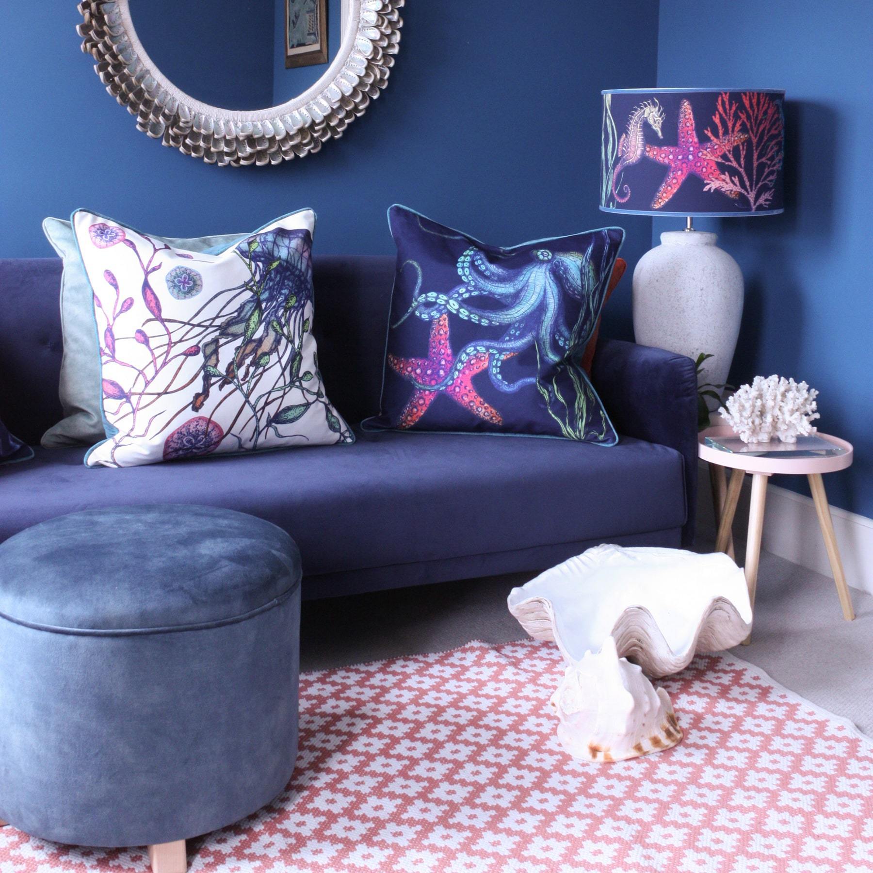 Rainbow Reef Navy Shade With Octopus,Seahorse,Starfish and Seaweed Design in bright colours on a white lampbase next to a navy sofa covered in our Reef cushions.Behind on the wall is a abalone mirror and in front of the sofa is a footstool and several shells on the floor. 