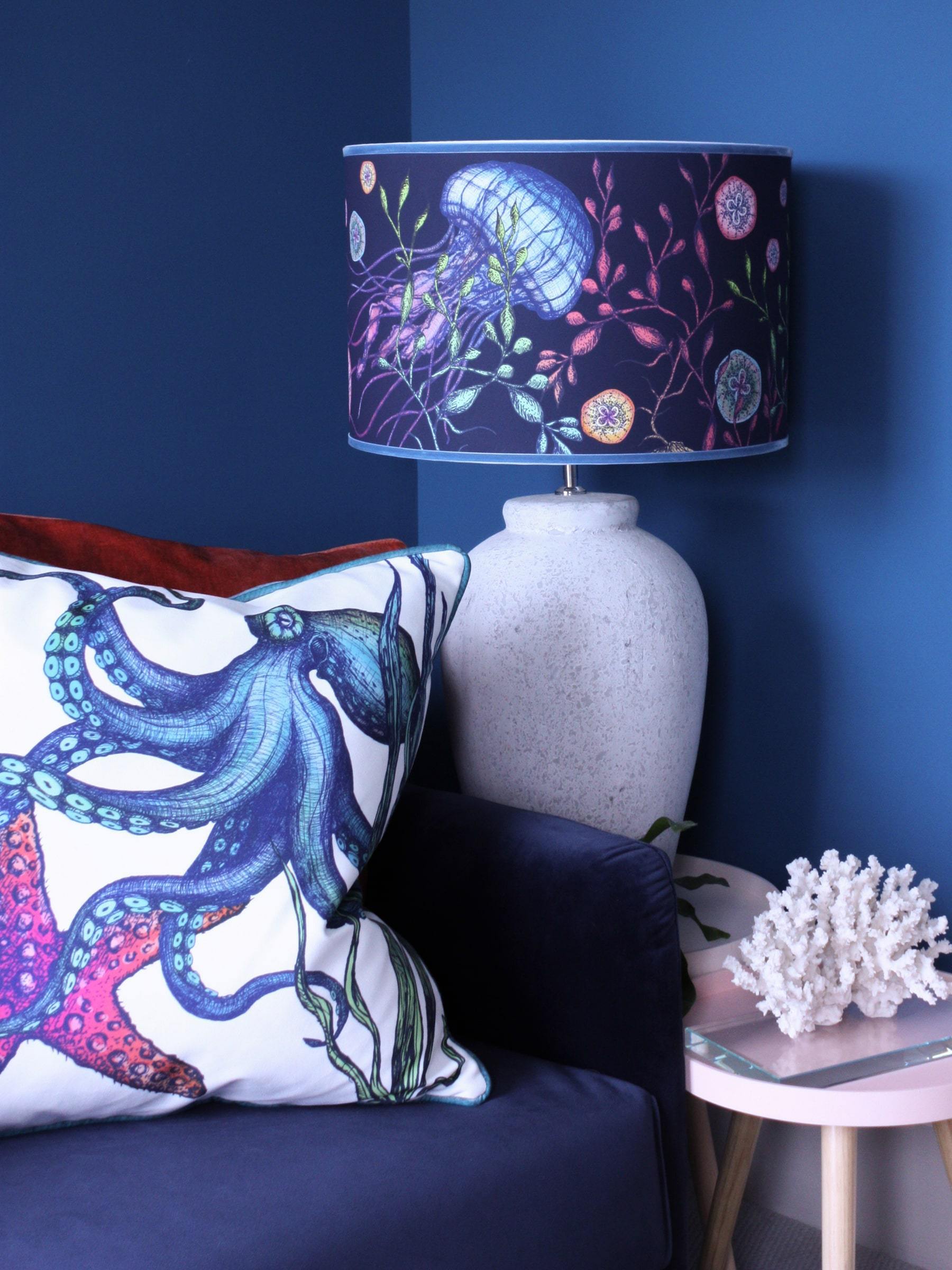 Canyons Reef Navy Shade With Jellyfish Design And Velvet Trim on a white lampbase on a table with a coral structure on it.This is next to a navy sofa with Reef cushions on