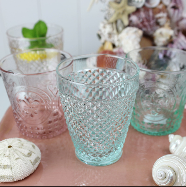 Aqua coloured Diamond cut glass tumbler on a table with other coloured glasses, in the background you can see a shell covered candlestick