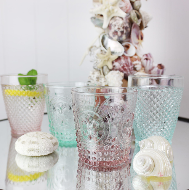Aqua coloured Diamond cut glass tumbler on a table with other coloured glasses, in the background you can see a shell covered candlestick and shells on a mirror tabletop