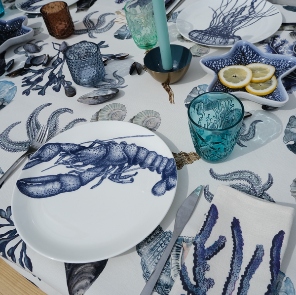 Coloured tumblers with embossed sea creatures in the glass,all set on a beachcomber tablecloth surrounded by place settings in our classic china range