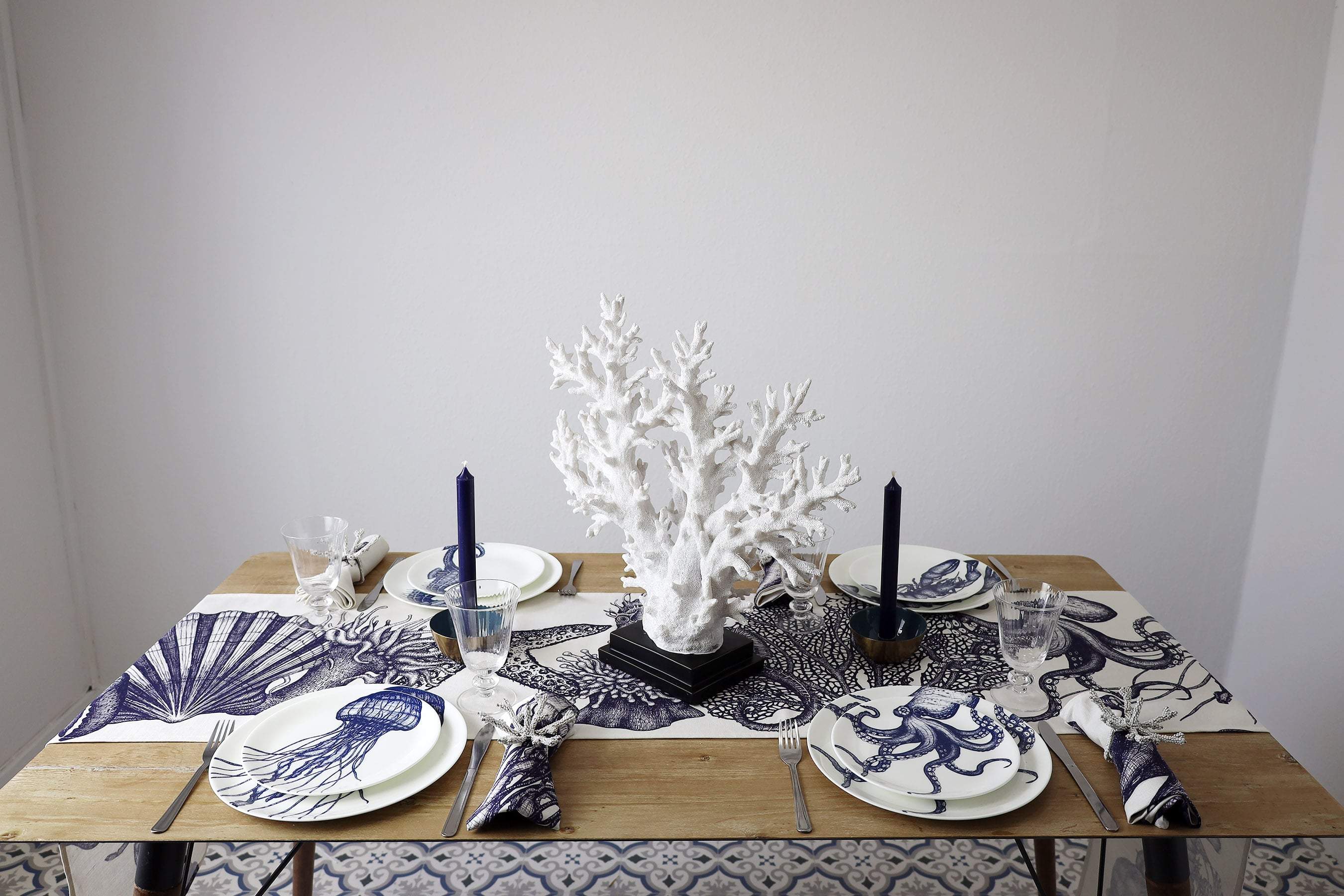 Blue & white linen napkins with seahorse, lobster, octopus and jellyfish set amongst a table setting.On the table is our tableware,table runner,cutlery,glasses and a large coral centrepiece 
