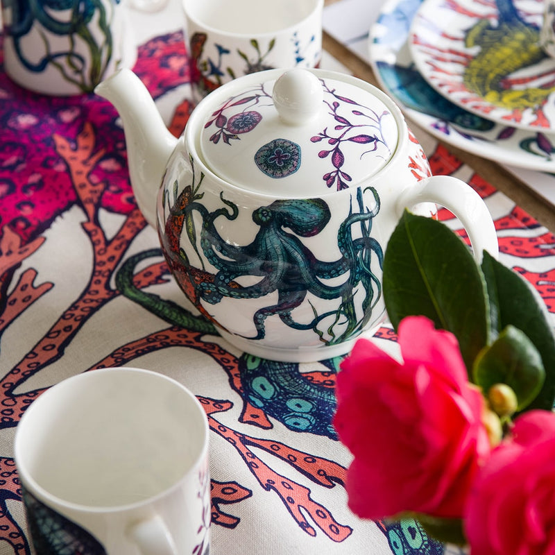 Bone China Teapot with hand drawn design in our Reef range showing the blue octopus and Starfish amongst the seaweed on a reef table runner featuring the same design.In the background are Reef plates and mugs and in the foreground is a pink camelia and another mug