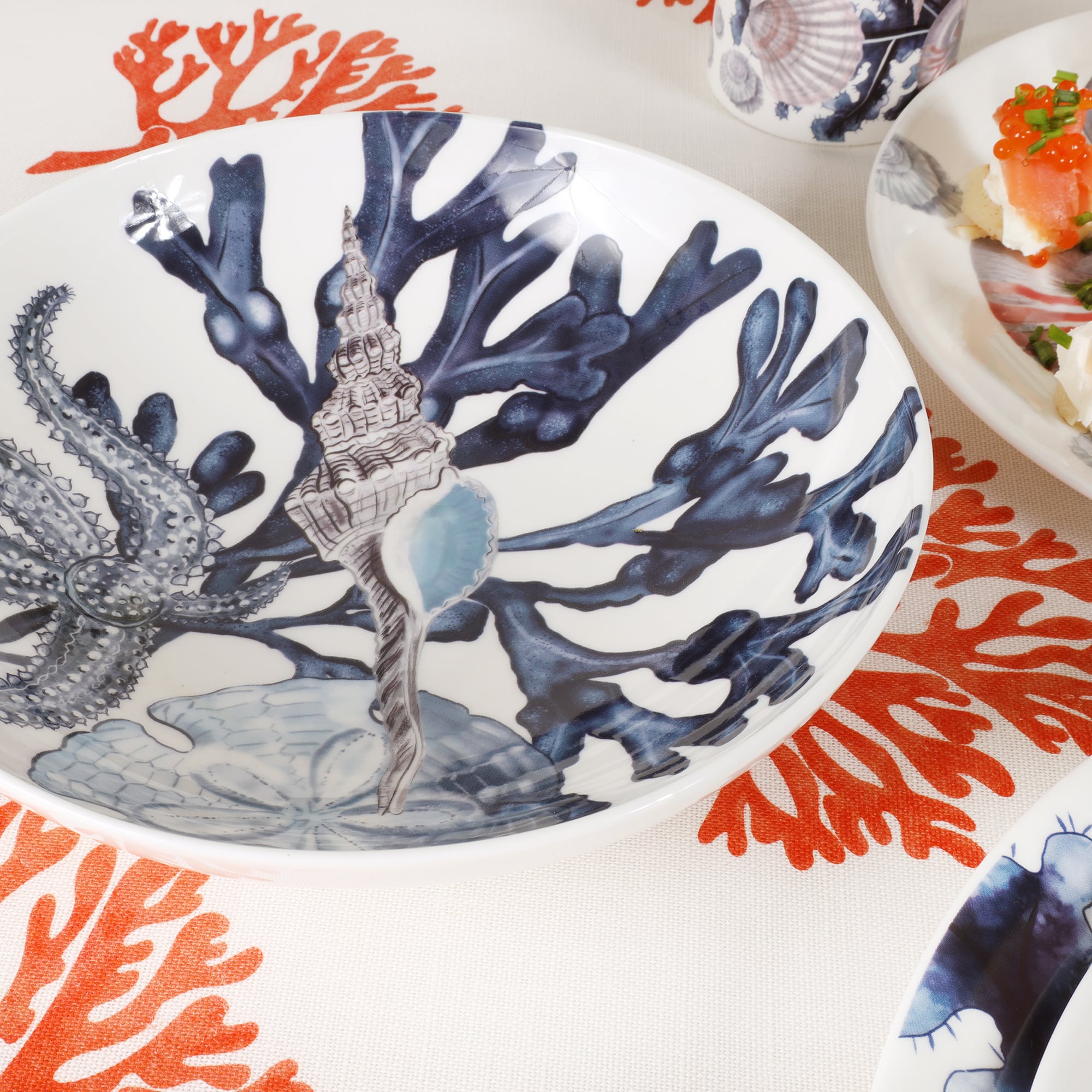 Close up of Beachcomber design pasta bowl has Dark Blue seaweed fronds,starfish,whelk and a sand dollar all in co ordinating blues.The bowl is placed on orange and white fistral fabric.In the background is a beachcomber jug and a plate of food