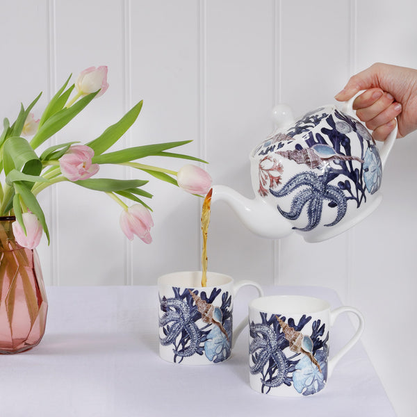 Bone China Teapot with hand drawn design in our beachcomber range with Seaweed,Starfish,Shells,Coral in mainly blues pouring tea into a matching mug.Also on the table is another mug of the same design and a pink vase with pink tulips
