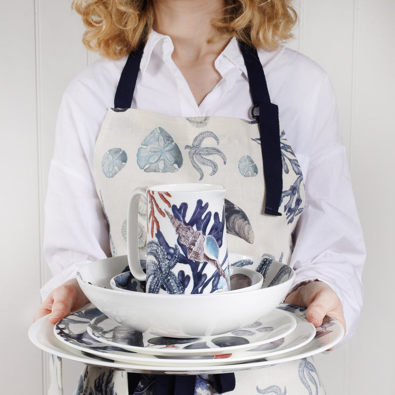 In the picture you can see a woman holding Beachcomber Extra Large Dinner plate,Dinner plate,side plate,pasta bowl,small bowl and a medium jug.The woman holding them is wearing a beachcomber apron over a white shirt.