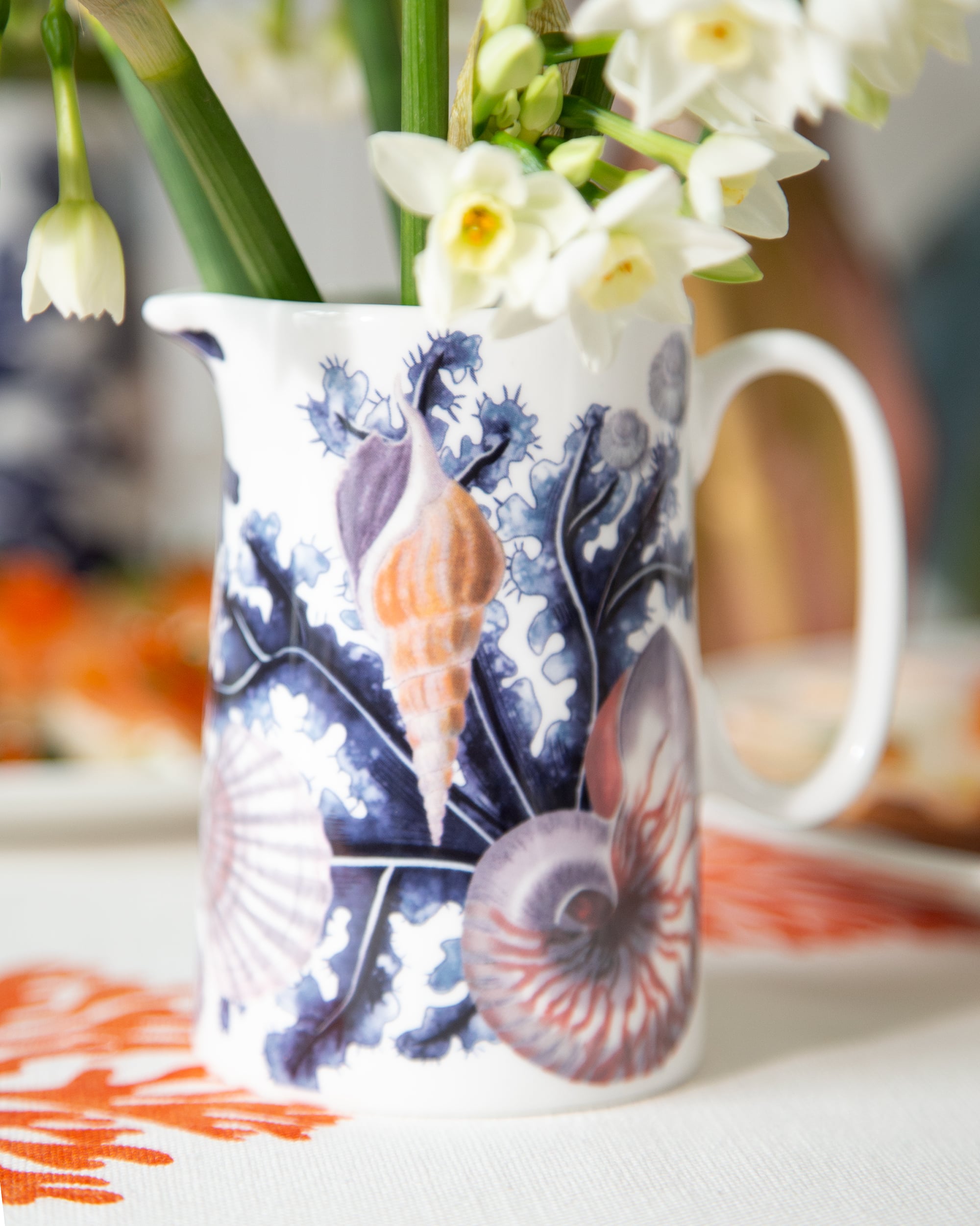 Close up of the Small size beachcomber jug showing blue seaweed, shells, scallops and a nautilus shell.In the jug are white daffodils with table decor in the background