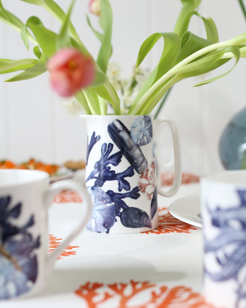 Bone china hand illustrated Medium size beachcomber jug showing shades of blue shells,seaweed and a starfish.In the jug are tulips in the vase on a fistral orange coral tablecloth
