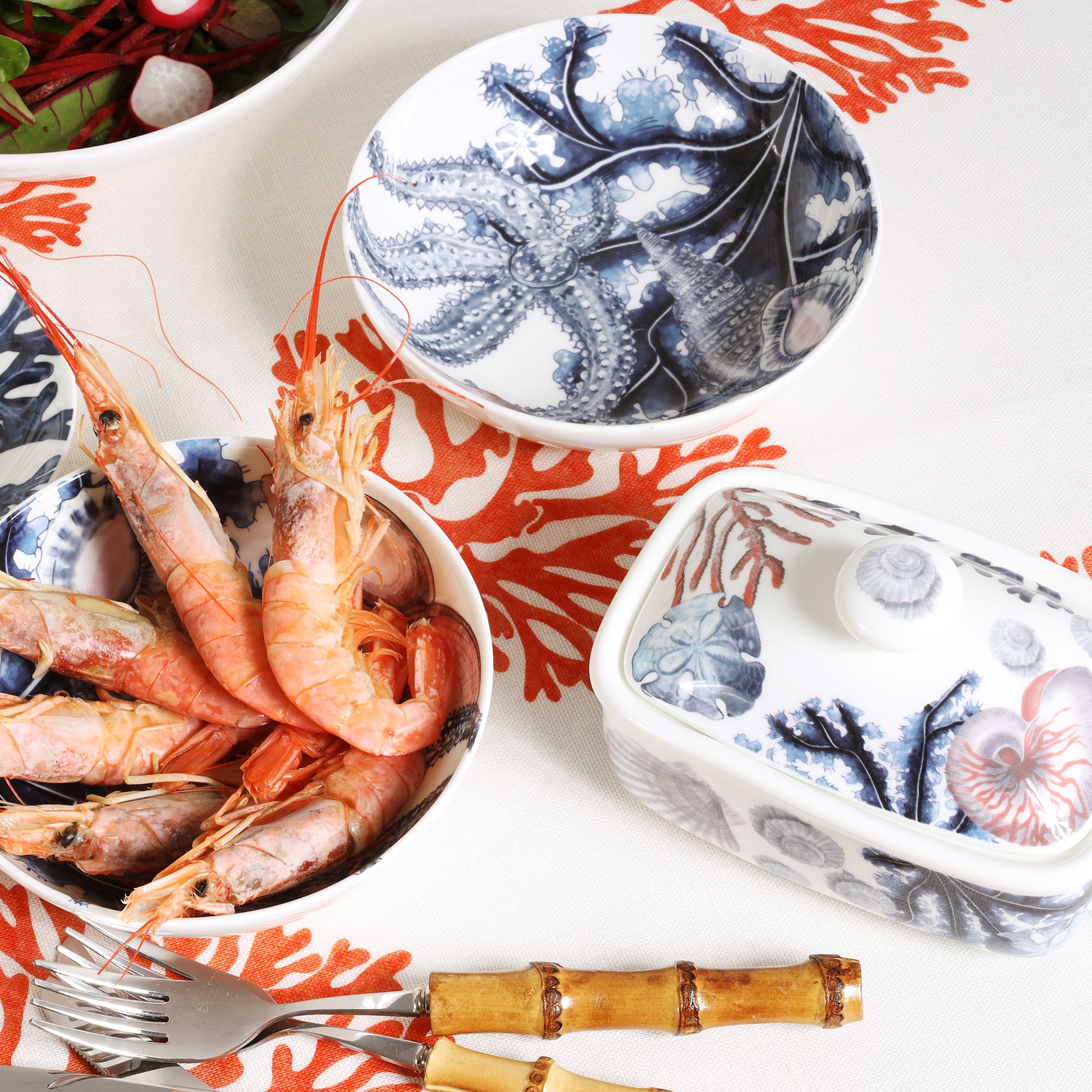 Butter dish in our Beachcomber range,with shells,seaweed and other sea themed designs all over the base and the lid.Also on the table are a couple of matching beachcomber bowls on a table cloth.In the bowls are prawns and a salad