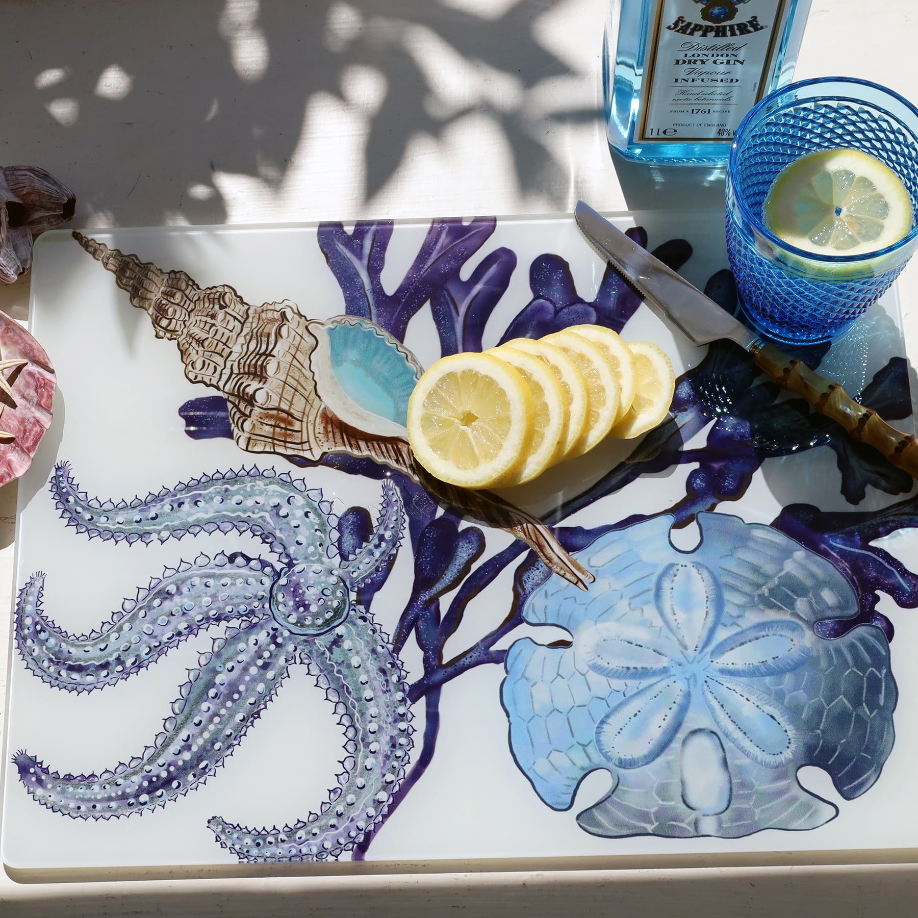 Glass worktop saver in our Beachcomber design with Starfish,a whelk,seaweed and a sand dollar design,placed on a table.On the worktop saver are sliced lemons,a knife and a glass of gin with the bottle behind it.