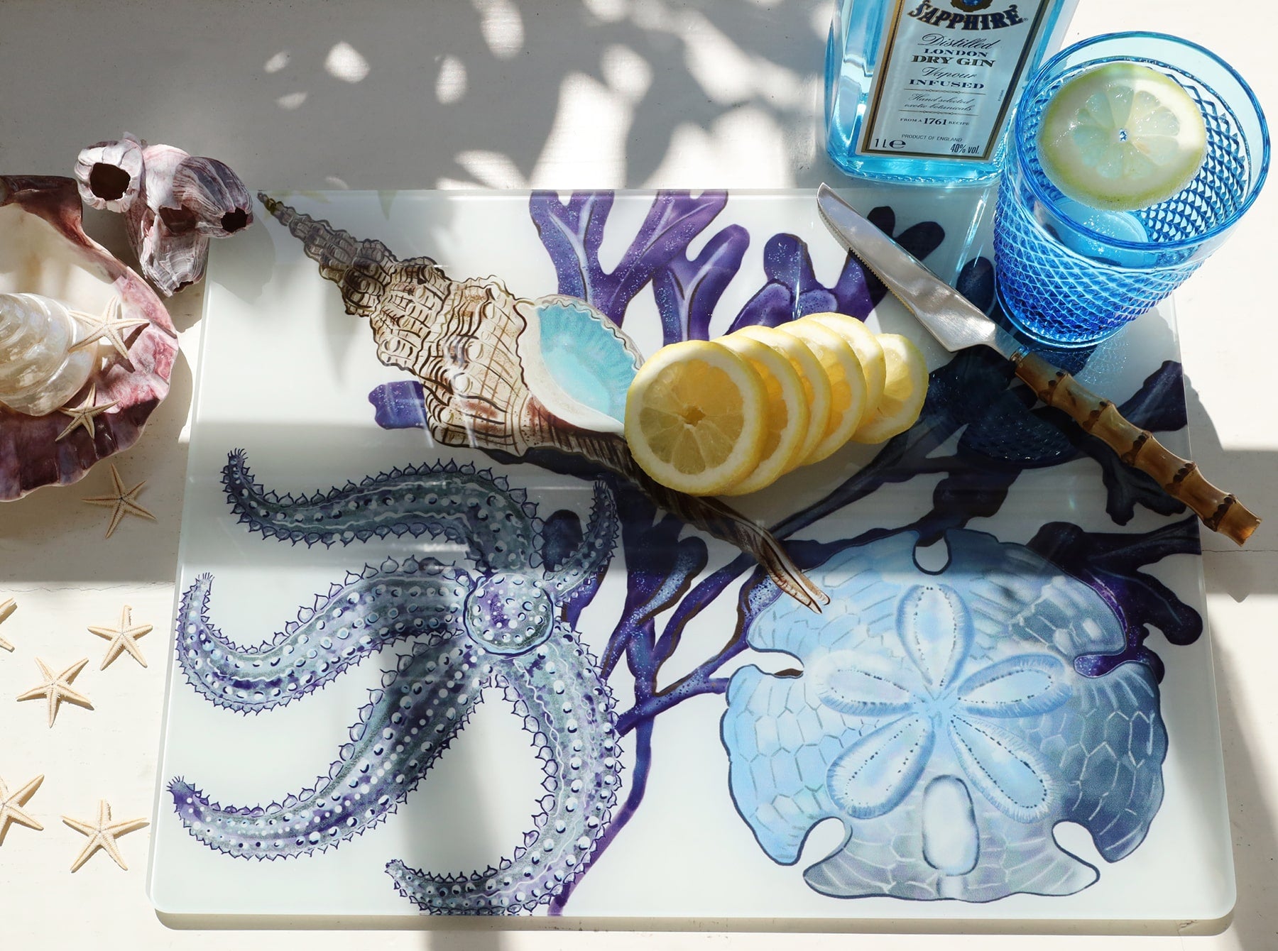 Glass worktop saver in our Beachcomber design with Starfish,a whelk,seaweed and a sand dollar design,placed on a table.On the worktop saver are sliced lemons,a knife and a glass of gin with the bottle behind it.