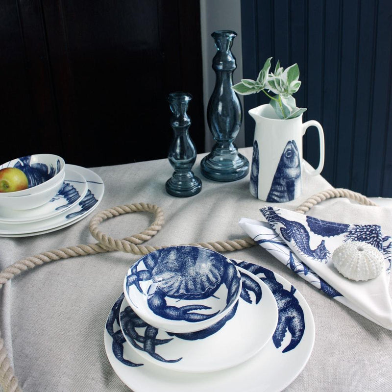 Bowl in Bone China in our Classic range in Navy and white in the Crab design stacked on a matching side plate and dinner plate placed on a tablecloth.On the table is a stack of lobster tableware,matching napkins,a mackerel jug and a couple of glass candle holders