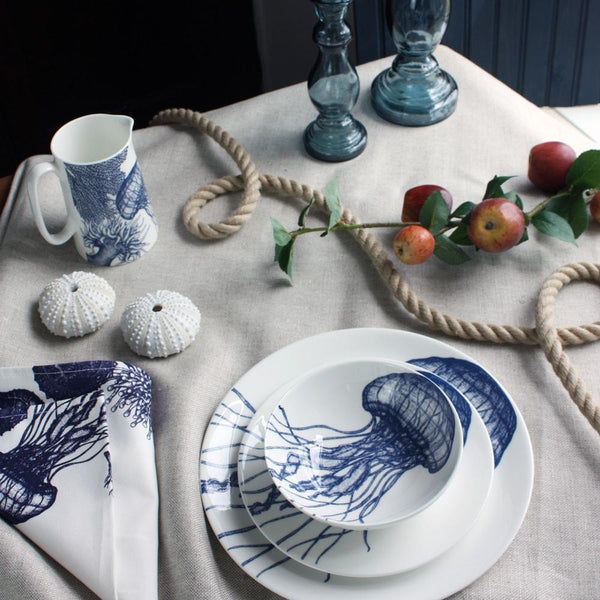 Bowl in Bone China in our Classic range in Navy and white in the Jellyfish design stacked on a matching side plate and dinner plate placed on a tablecloth.On the table is a matching napkins,a mackerel jug,a rope winding on the table and a couple of glass candle holders