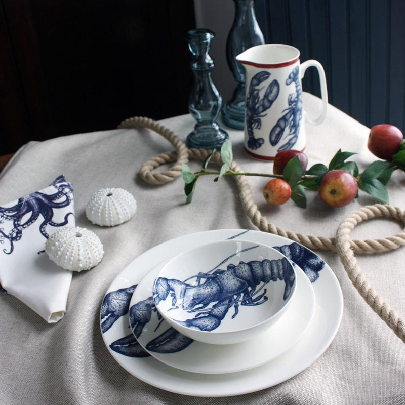 Bowl in Bone China in our Classic range in Navy and white in the Lobster design stacked on a matching side plate and dinner plate placed on a tablecloth.On the table is a matching napkins,a lobster jug and a couple of glass candle holders and an apple branch