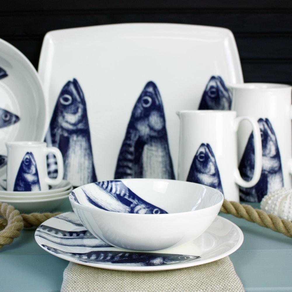 Bowl in Bone China in our Classic range in Navy and white in the Mackerel design stacked on a matching side plate and dinner plate placed on a tablecloth.On the table is a mackerel jug,a rope winding on the table.In the background are more matching tableware including a large platter 