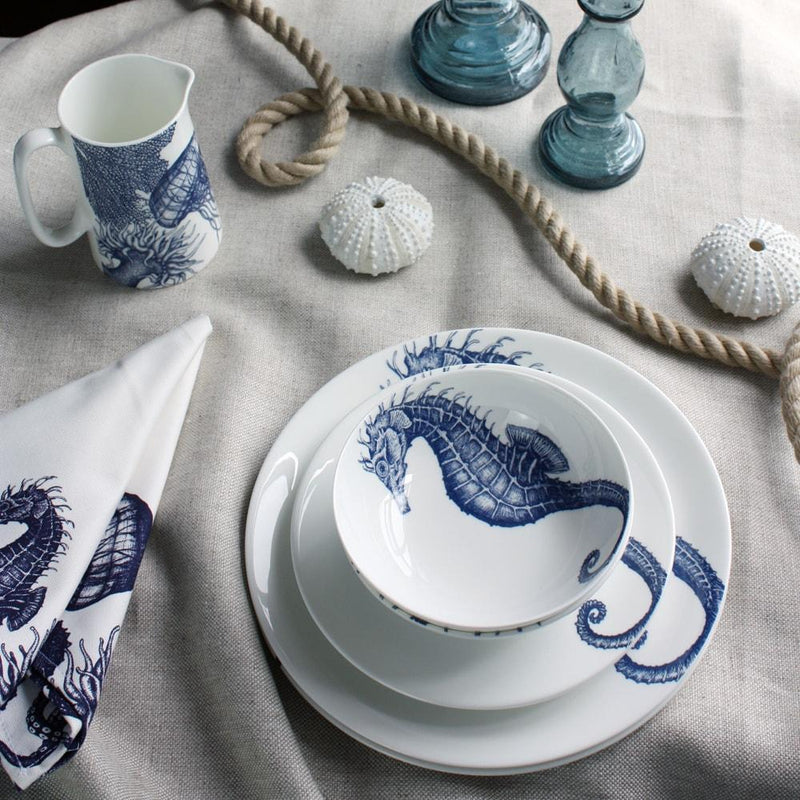 Bowl in Bone China in our Classic range in Navy and white in the Seahorse design stacked on a matching side plate and dinner plate placed on a tablecloth.On the table is a matching napkins,a seahorse jug,a rope winding on the table and a couple of glass candle holders