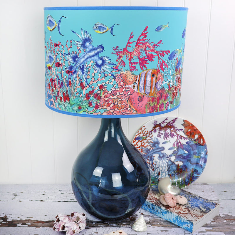 Coral Bay design lampshades in Turquoise with blue trim on a petrol glass lampbase placed on a table.Next to the lampbase is a Coral Ray plate behind a book and some shells 